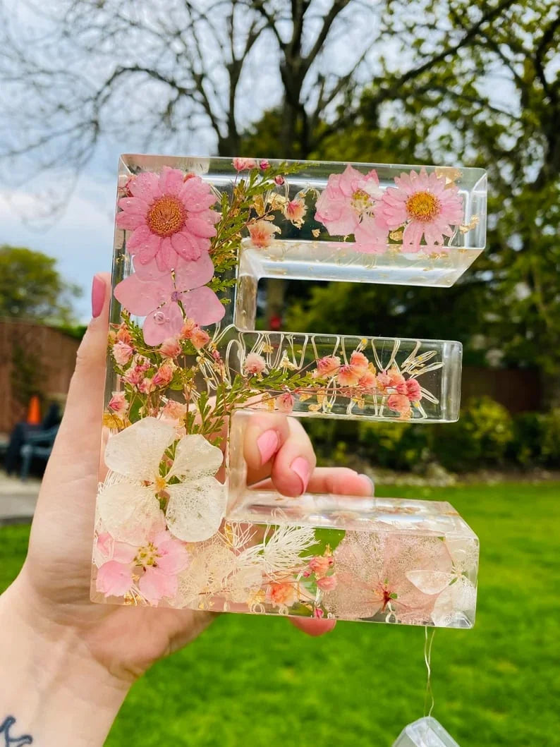 Handgjord bokstavsformad harts-nattlampa med rosa blommor som hålls i handen utomhus.