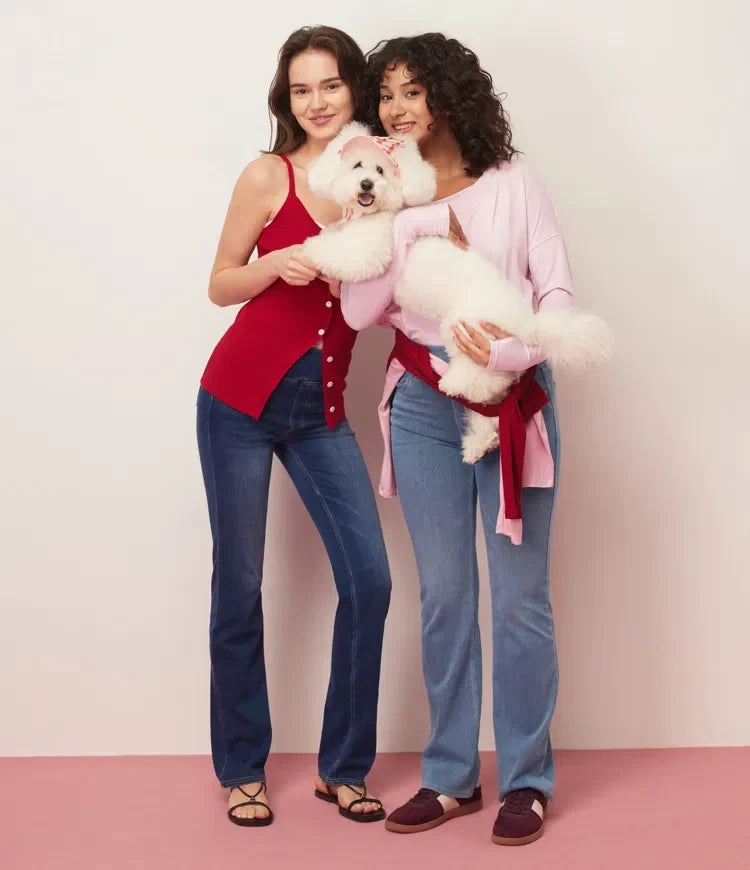 Två glada kvinnor som visar upp blå bootcut jeans tillsammans med en hund