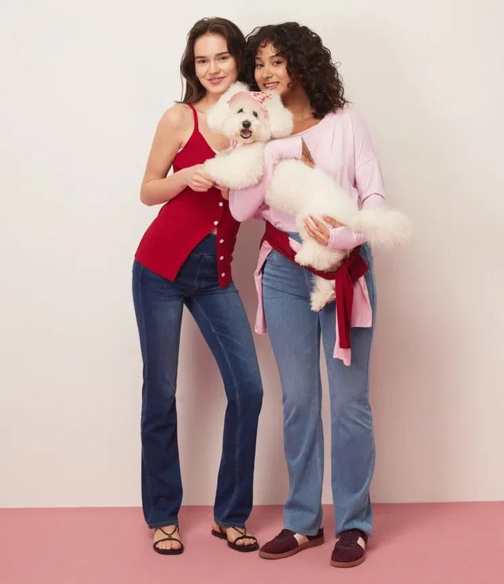Två glada kvinnor som visar upp blå bootcut jeans tillsammans med en hund