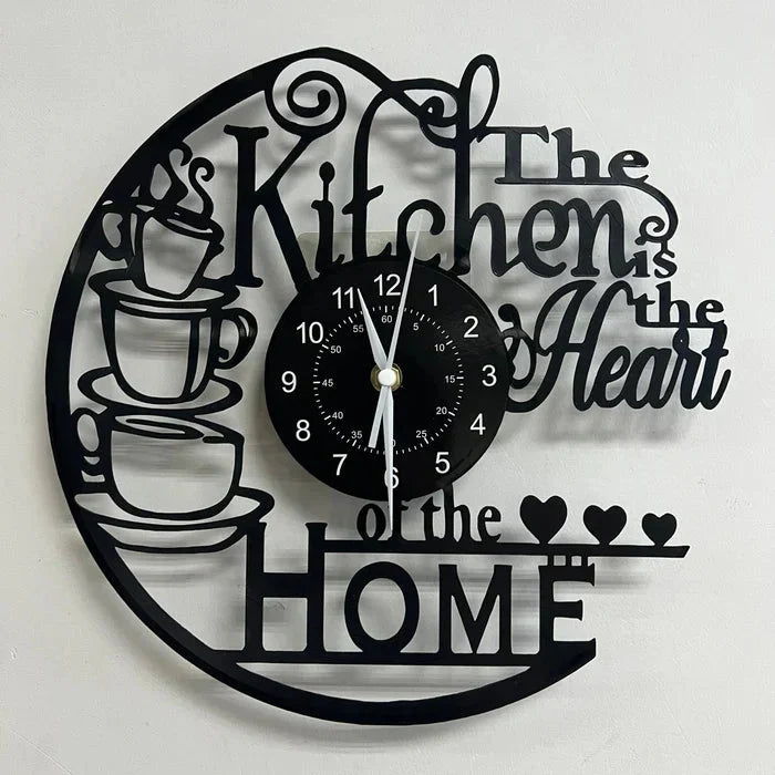 Svart väggklocka med kaffemotiv och texten The Kitchen is the Heart of the Home på en vit vägg