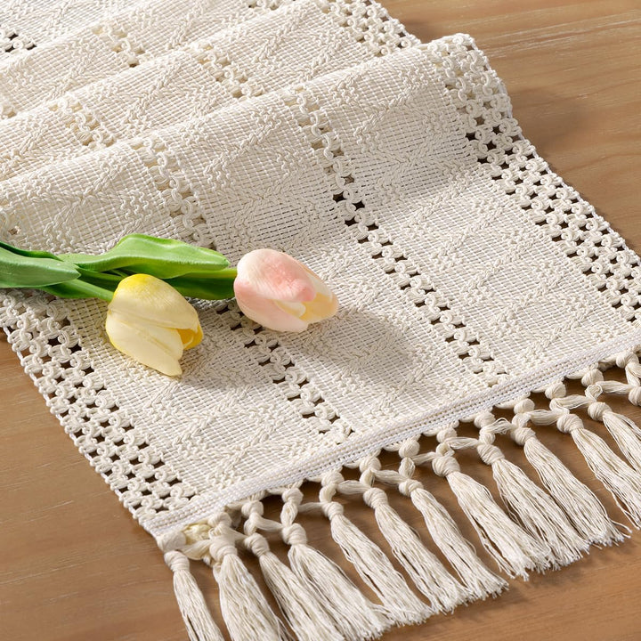 Närbild på en beige makramébordslöpare med handknutna tofsar och vårblommor på träbord
