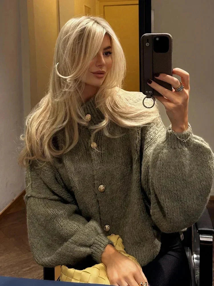 Blond kvinna i mörkgrön chunky cardigan med guldknappar som tar spegeltoalett