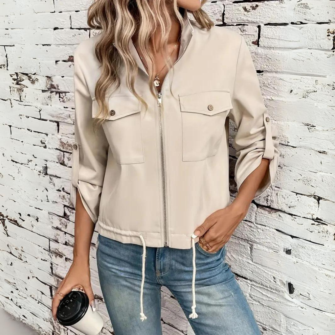 Beige damjacka med dragkedja och påsydda fickor bodd mot vit tegelvägg