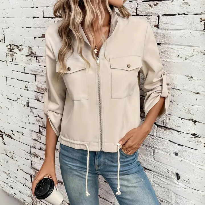 Beige damjacka med dragkedja och påsydda fickor bodd mot vit tegelvägg
