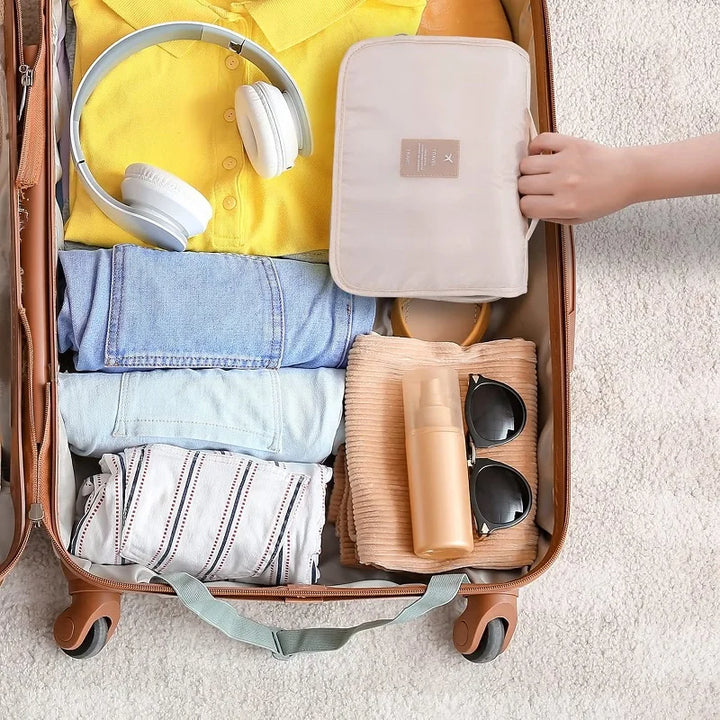 Beige hängande necessär som packas ner i en öppen resväska tillsammans med kläder och hörlurar