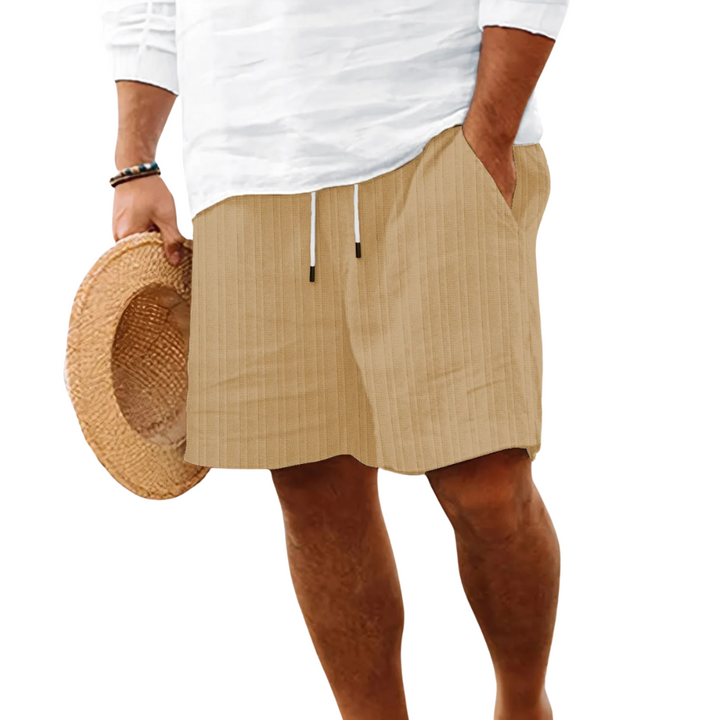 Man iklädd beigea texturerade herrshorts med matchande vit skjorta och stråhatt vid stranden