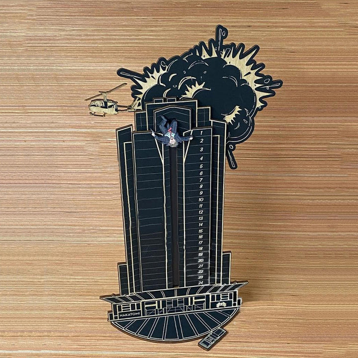 "Detaljerad trä Adventskalender med Die Hard-motiv, visar Hans Gruber faller från brunblå Nakatomi Plaza, roterbar design."