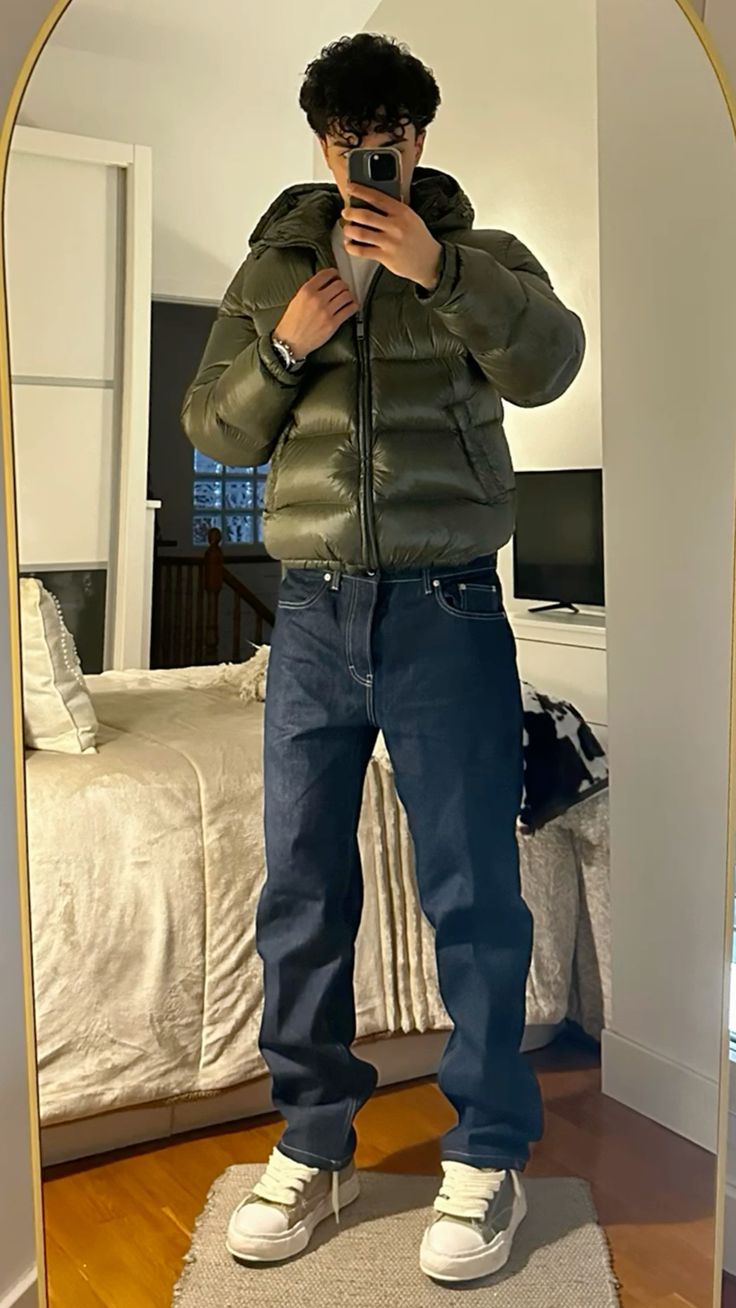 Man iklädd grön quiltad jacka med huva och mörka jeans framför spegeln
