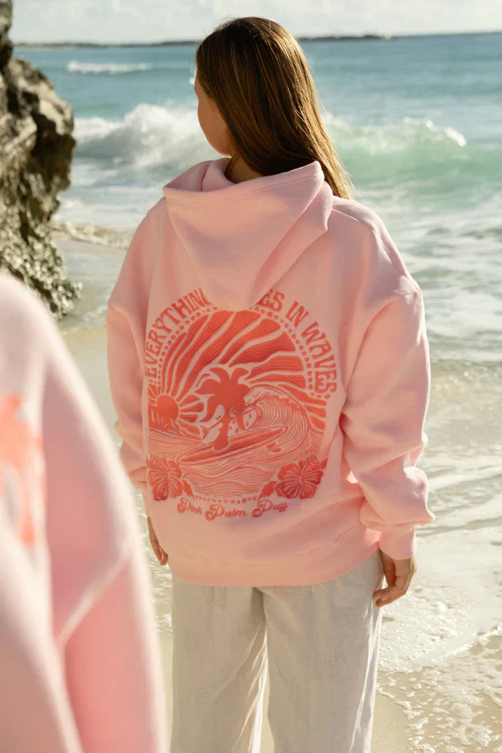 Kvinna i rosa hoodie med tropiskt palm- och vågtryck på ryggen vid havet